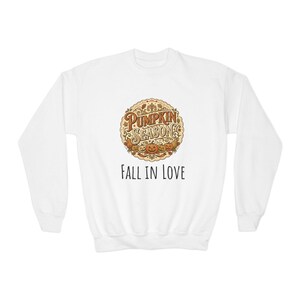 Könnte beinhalten: Ein weißes Langarm-Sweatshirt mit einer kreisförmigen Grafik, die den Text PUMPKIN SEASON in braunen Buchstaben zeigt, umgeben von orangefarbenen Kürbissen und Herbstblättern. Unter der Grafik steht der Text FALL IN LOVE in Schwarz.