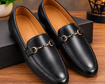 Könnte beinhalten: Ein Paar schwarze Leder-Loafer mit einem goldfarbenen Metall-Trensendetail über dem Vorderblatt. Die Schuhe haben ein glattes schwarzes Äußeres, eine abgerundete Zehenpartie und ein beigefarbenes Innenfutter im klassischen Slipper-Design.