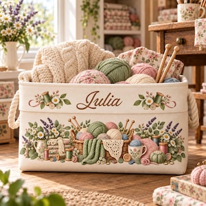 May include: A rectangular off-white fabric storage basket with rope handles, featuring the name Julia in brown script, decorated with illustrations of yarn, knitting needles, flowers, and sewing supplies, filled with various yarn balls.