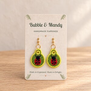 May include: A pair of embroidered ladybird earrings on a display card labelled Bubble & Mandy Handmade Earrings. The earrings feature a red and black ladybird on a bright green circular background with gold-toned hooks. The card text reads Made in Gippsland. Made to Delight.