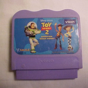 May include: Purple VTech V.Smile cartridge for the Disney Pixar Toy Story 2 game. The cartridge features images of Buzz Lightyear, Woody, and Jessie. The text on the cartridge reads "Operation: Rescue Woody!"