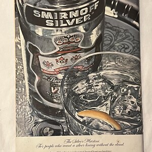 May include: A vintage advert for Smirnoff Silver vodka. The image shows a bottle of Smirnoff Silver, a glass of ice water with a lemon peel, and text that reads "The Silver Martini." The bottle has a silver label and a crown.