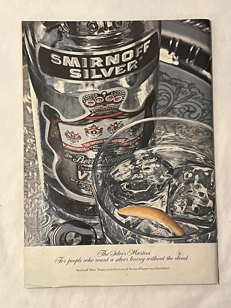 May include: A vintage advert for Smirnoff Silver vodka. The image shows a bottle of Smirnoff Silver, a glass of ice water with a lemon peel, and text that reads "The Silver Martini." The bottle has a silver label and a crown.
