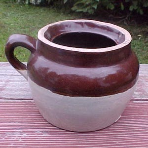 May include: A brown and white glazed stoneware crock with a handle. The crock has a wide mouth and a rounded body.