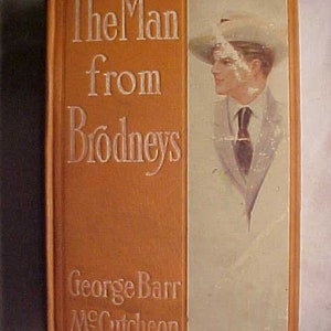 May include: An orange book with the title "The Man from Brodneys" by George Barr McCutcheon. The cover features a black and white illustration of a man wearing a hat and suit.