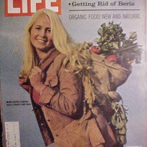 May include: LIFE magazine cover featuring a smiling woman with blonde hair, wearing a brown jacket, and carrying a bundle of vegetables. The magazine title "LIFE" is in large white letters. Text includes "KHRUSHCHEV REMEMBERS" and "ORGANIC FOOD: NEW AND NATURAL."
