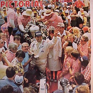 May include: A group of people gathered around a trophy, many wearing racing jackets and hats. The image is from a magazine called "Racing Pictorial".
