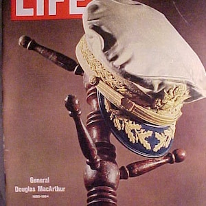 May include: LIFE magazine cover featuring a portrait of General Douglas MacArthur. The cover shows a military hat with gold trim and a blue band, resting on a wooden hat stand. The magazine's title is in large white letters on a red background.