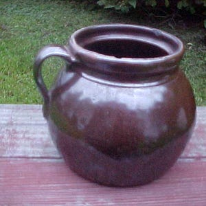May include: A brown glazed ceramic crock with a single handle. The crock has a rounded body and a wide opening.