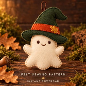 May include: A cream-coloured felt ghost ornament wearing a dark green witch hat with an orange band and a small yellow oak leaf accent. The ghost has brown embroidered eyes and a smile, with a brown hanging loop at the top of the hat.