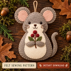 May include: A felt mouse ornament featuring a grey body with a cream-coloured belly and inner ears, holding a sprig of three red berries and two green leaves. The mouse has black eyes, a stitched nose, and a jute loop for hanging at the top.