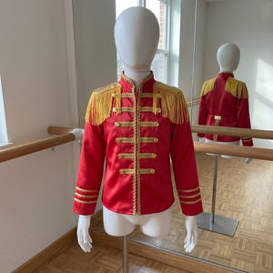 May include: A bright red jacket featuring gold braided trim, a high collar, and long sleeves. The shoulders are adorned with gold fringe epaulettes. The front closure includes five rows of gold buttons and decorative gold cord detailing.