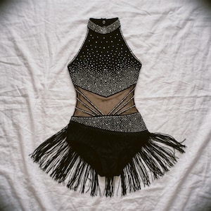 May include: A black leotard featuring a high neckline and sleeveless cut, adorned with numerous silver rhinestones. The garment includes sheer mesh side panels with diagonal straps and a bottom section finished with long black fringe trim.