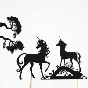 以下が含まれることがあります： 森のシーンが描かれたプラットフォームの上に立っている2頭のユニコーンの黒いシルエット。ユニコーンは異なる方向を見ており、角と花を頭に付けています。