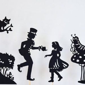 May include: Black silhouette cutouts of characters from Alice in Wonderland, including the Mad Hatter, Alice, and the Cheshire Cat. The characters are posed in a whimsical scene, perfect for shadow puppetry.