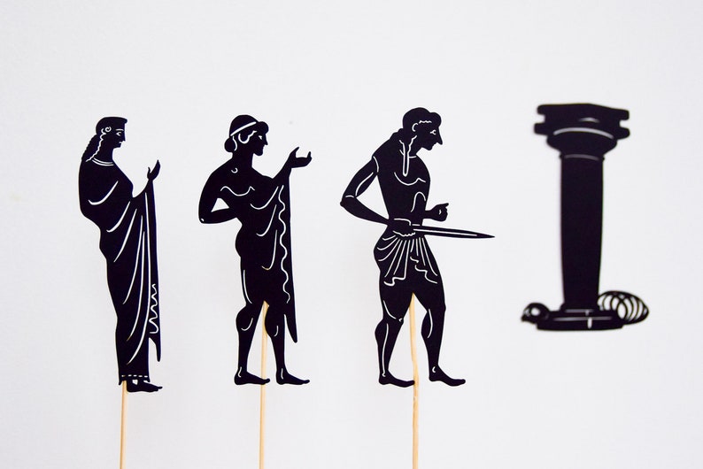 May include: Black paper cutouts of ancient Greek figures and a column on wooden sticks. The figures include a woman in a draped gown, a man with a draped cloth, a man holding a sword, and a classical column.