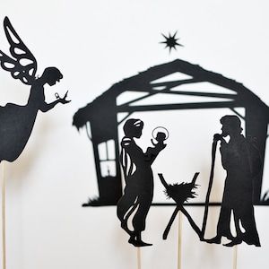 May include: Black paper silhouette figures of a nativity scene with a star above the stable, Mary holding baby Jesus, Joseph holding a staff, and an angel with wings.
