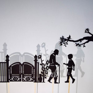 May include: Black silhouette shadow puppets of two figures, an older person with a cane and a younger person, standing in front of a gate with a decorative design. The figures are in front of a white background.