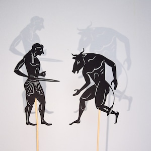 May include: Black paper cutouts depicting a mythological scene. A figure with a sword faces a bull-headed humanoid. Shadows of the figures are visible on the white background. The figures are mounted on wooden sticks.