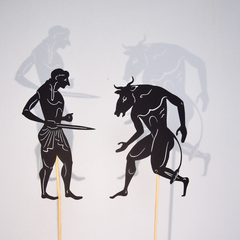 May include: Black paper cutouts depicting a mythological scene. A figure with a sword faces a bull-headed humanoid. Shadows of the figures are visible on the white background. The figures are mounted on wooden sticks.