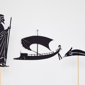 May include: Black paper cutouts of a man, a sailboat, and a dolphin on wooden sticks. The man is in profile, holding a staff. The sailboat has a large sail. The dolphin is leaping. All are against a white background.