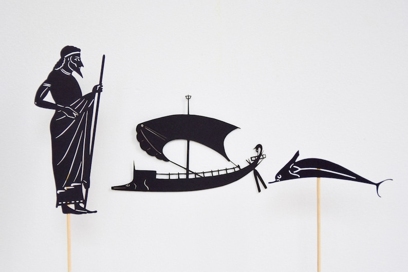May include: Black paper cutouts of a man, a sailboat, and a dolphin on wooden sticks. The man is in profile, holding a staff. The sailboat has a large sail. The dolphin is leaping. All are against a white background.