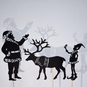 May include: Black silhouette cutouts of Santa Claus, a reindeer, and an elf. Santa is waving, the reindeer has antlers, and the elf is holding a small object.