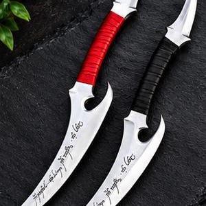 May include: Two curved daggers with silver-toned blades featuring engraved script. One dagger has a bright red wrapped handle, while the other has a black wrapped handle. Both blades have a distinct notch near the hilt and a sharp, pointed tip.