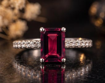 Puede incluir: Un anillo de tono plateado con una gran piedra preciosa rectangular de color rojo intenso engastada en cuatro garras. La banda está adornada con una hilera de pequeñas piedras claras de corte redondo a ambos lados de la piedra central.