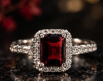 May include: A silver-toned ring featuring a rectangular emerald-cut deep red garnet center stone, surrounded by a halo of small clear round stones. The band is also set with small clear round stones along the sides of the ring.