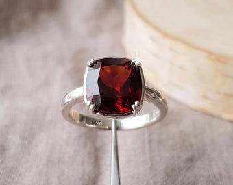 May include: A silver ring featuring a large, cushion-cut, deep red garnet gemstone held by four prongs. The inner band of the ring is clearly stamped with the number 925, indicating sterling silver composition. The setting is polished and smooth.