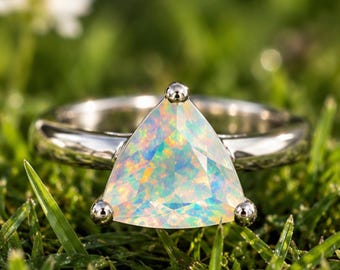 May include: A silver-toned ring featuring a large, triangular-cut opal gemstone. The stone displays a play of iridescent colors including pink, green, and blue. The opal is secured by three small, rounded metal prongs at each corner.