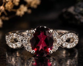 Puede incluir: Un anillo de plata con una gran piedra preciosa de color rojo intenso y corte ovalado en el centro, sujeta por cuatro garras. La banda está adornada con pequeñas piedras claras de corte redondo en un patrón de infinito a los lados.