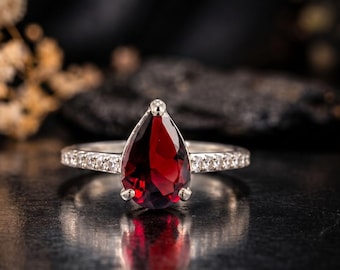 May include: A silver ring featuring a large, pear-shaped, deep red garnet gemstone held by three prongs. The band is accented with a row of small, clear, round-cut white stones embedded along both sides of the shank.