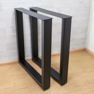 May include: A pair of matte black metal table legs with a rectangular frame design. Each leg features a flat top mounting plate with pre-drilled holes for attachment, constructed from sturdy square tubing in a vertical orientation.