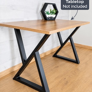 May include: A pair of black metal table legs featuring a geometric, angular design that supports a wooden tabletop. A dark gray sign with white text reads Tabletop Not included, with an arrow pointing to the wooden surface.