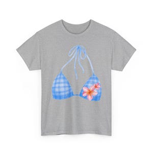 Peut inclure: Un t-shirt gris chiné arborant un motif de haut de bikini à carreaux vichy bleu clair et blanc. L'illustration du haut de bikini présente de fines bretelles bleues et un groupe de fleurs tropicales roses et orange sur le bonnet droit.