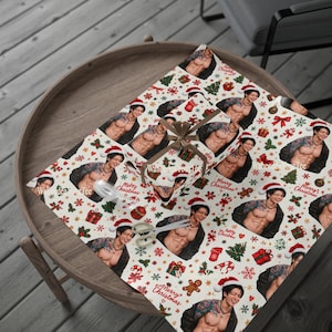May include: Wrapping paper featuring a repeating pattern of a shirtless man with tattoos wearing a Santa hat, interspersed with Christmas trees, gifts, candy canes, and the text Merry Christmas. A pair of scissors rests on the paper.