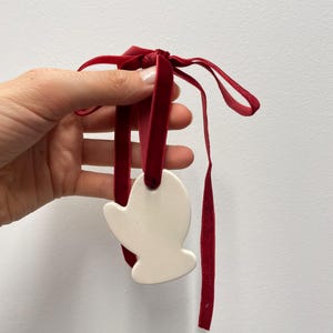 Peut inclure: Un ornement en céramique blanc cassé lisse en forme de moufle, doté d'un trou sur le dessus. Un ruban de velours rouge profond est passé dans le trou et noué en boucle, avec deux longs pans de ruban qui pendent sous l'ornement.