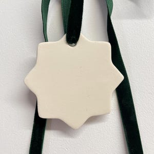 Peut inclure: Un ornement en forme d'étoile à huit branches de couleur blanc cassé, fabriqué dans un matériau lisse et mat. L'ornement présente un trou unique au sommet, dans lequel un ruban de velours vert forêt foncé est passé et noué en boucle.