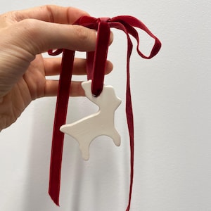 Peut inclure: Un ornement en céramique blanche en forme de silhouette de renne, suspendu à un ruban de velours rouge foncé noué en boucle. La figurine de renne à la finition mate et lisse présente une forme simple avec un trou au sommet pour le ruban.
