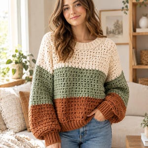 Puede incluir: Un suéter de ganchillo de manga larga con un diseño de bloques de color con rayas horizontales en crema, verde salvia y naranja óxido. La prenda tiene un cuello redondo acanalado, puños acanalados y un dobladillo acanalado con textura.
