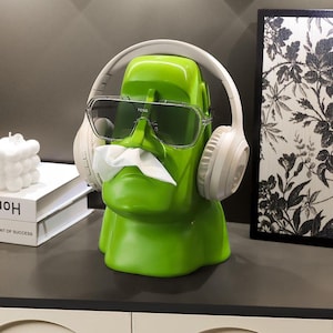 May include: A bright green Moai-shaped tissue dispenser featuring a stylised head design. The figure wears large, clear-framed sunglasses with the text FENG printed on the bridge and white over-ear headphones. A white tissue protrudes from the nose area.