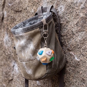 Puede incluir: Un llavero beige con forma de dodecaedro que presenta pequeñas presas de escalada de varios colores en cada cara. El llavero cuelga de un mosquetón y una anilla de metal plateado, sujetos al borde de una bolsa de magnesio de color verde oliva.