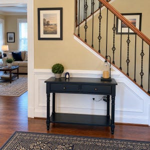 May include: A black rectangular console table with two drawers, turned legs, and a lower shelf. The table surface holds a small green potted plant, a small penguin figurine, and a tall glass candle lamp with a black metal base.