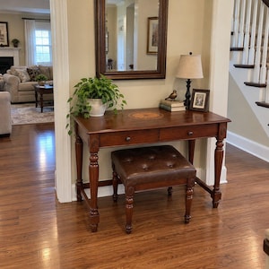 May include: A dark wood writing desk with two drawers, turned legs, and a circular inlay on the top surface. A brown tufted leather ottoman is tucked underneath. On the desk sits a potted green plant, a lamp, two books, and a small bird figurine.
