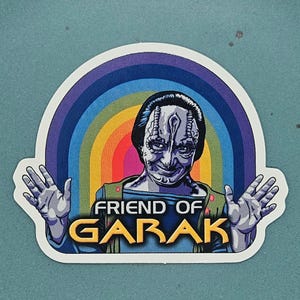 Op de afbeelding: Een sticker met een gestileerd portret van Garak uit Star Trek, met een regenboogachtergrond. De tekst "FRIEND OF GARAK" is vetgedrukt en geel weergegeven.