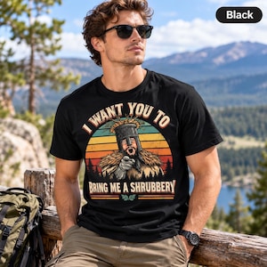 May include: A black short-sleeved t-shirt featuring a graphic of a knight in a helmet pointing forward. The text reads I WANT YOU TO BRING ME A SHRUBBERY. The design includes a sunset background with horizontal stripes and forest silhouettes.