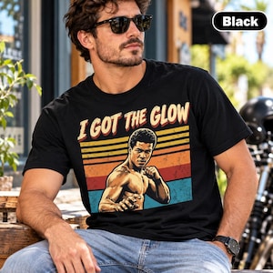 May include: A black short-sleeve t-shirt featuring a graphic print of a martial artist in a fighting stance. The design includes the text I GOT THE GLOW above the figure, set against a background of horizontal stripes in orange, yellow, and blue.