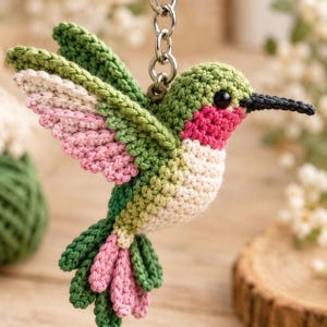 Puede incluir: Un llavero de colibrí tejido a ganchillo con hilo verde, crema y rosa. El pájaro tiene un pico negro, un pequeño ojo negro y está unido a una anilla de metal plateado. Las alas están extendidas, mostrando el patrón de colores en capas.